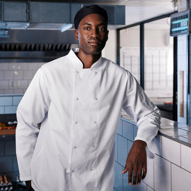 A chef wearing a long-sleeved standing in a kitchen leaning on a counter facing forward